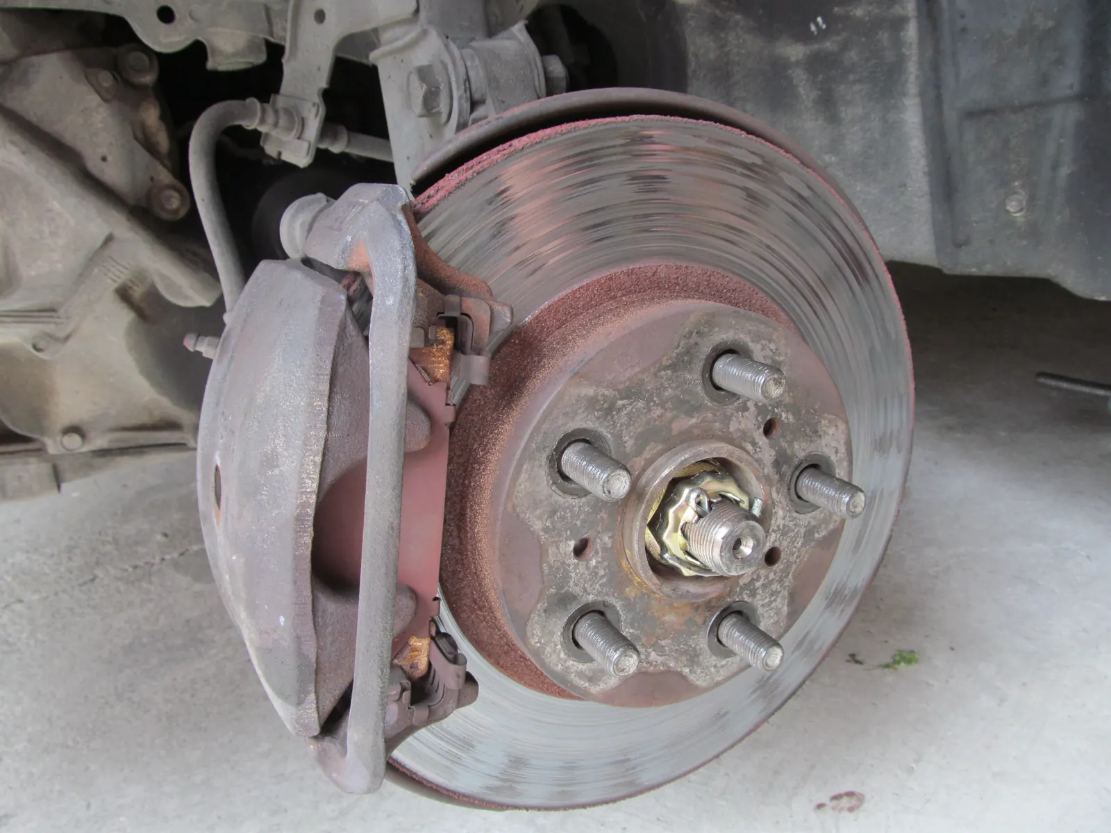 Worn passenger-car brake disc with concentric grooves, rusted hat, and a grey caliper, wheel off