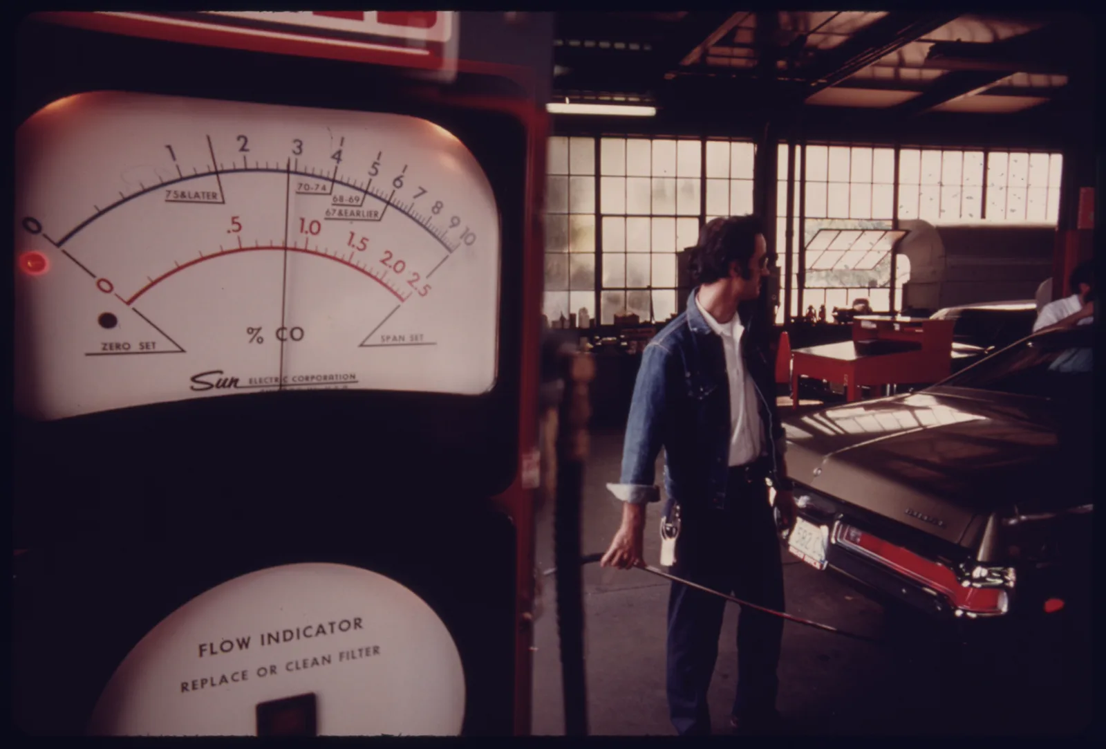 Sun exhaust analyzer CO gauge in the foreground, inspector holding a sample hose beside a black sedan