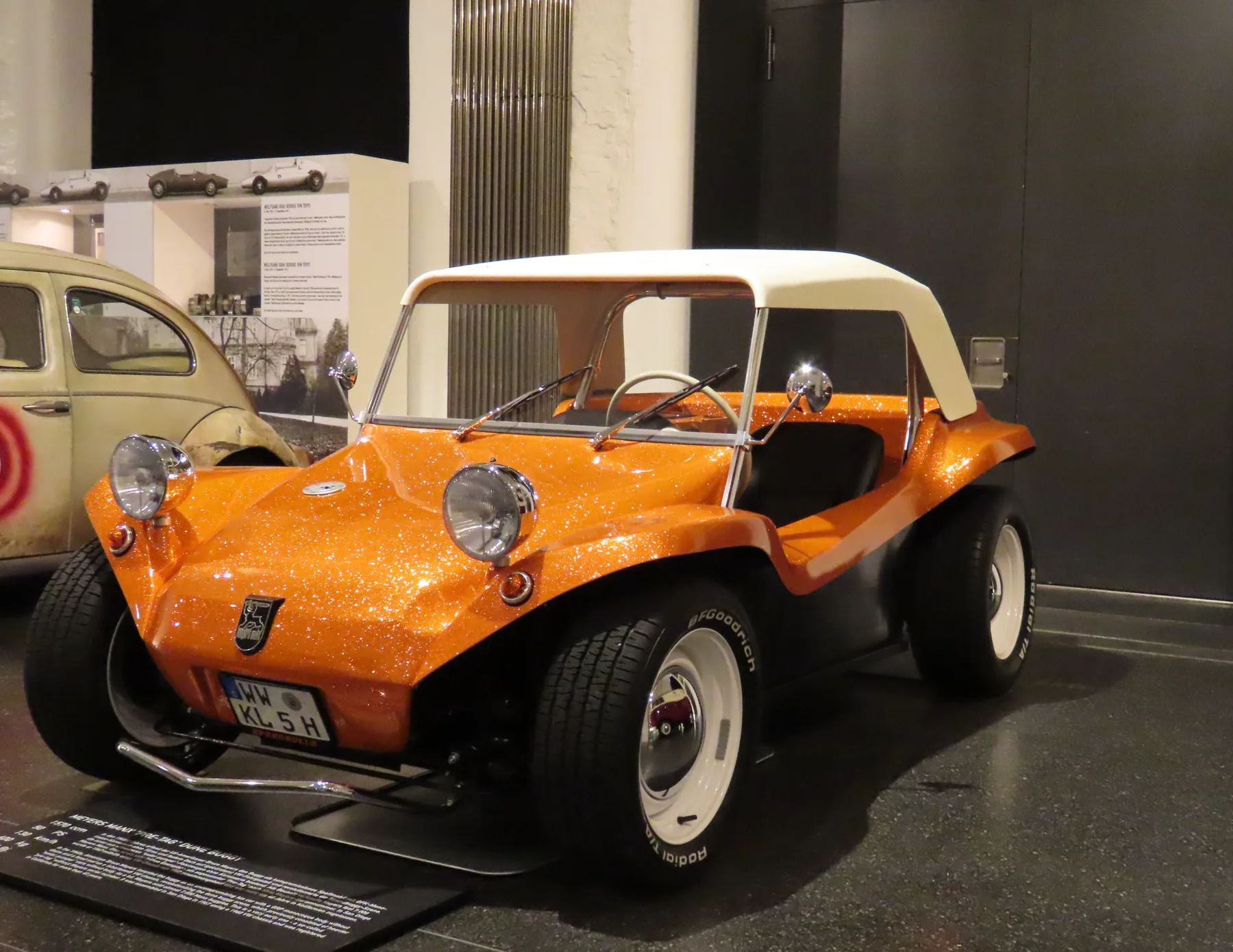 Orange metal-flake dune buggy with a white top and chrome headlamps, in a museum beside a Volkswagen Beetle