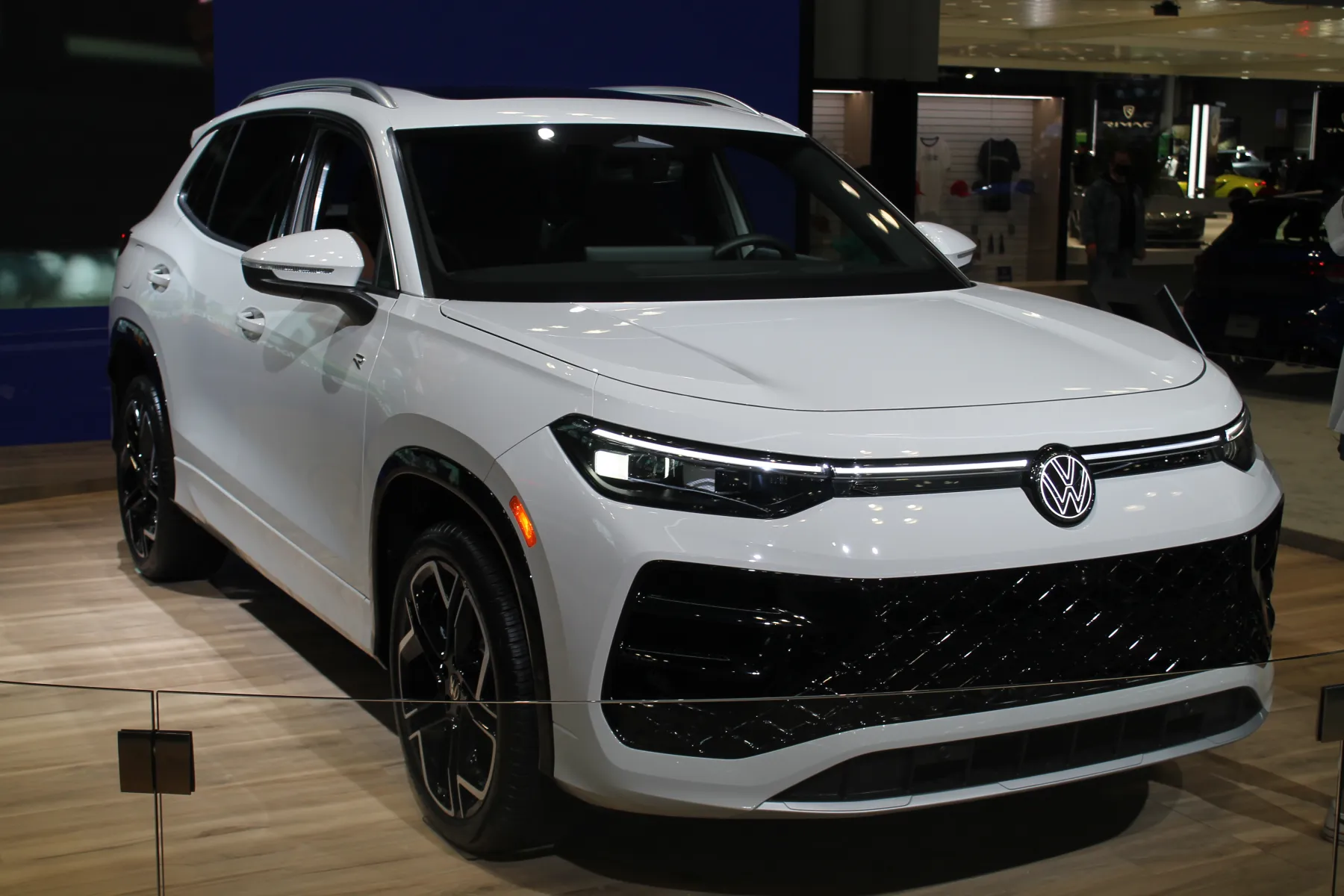 Front three-quarter of a white Volkswagen Tiguan on a light wood floor at an auto show, nose to the right, with a rope barrier across the foreground. A bright lamp bar runs across the nose through an illuminated VW roundel. The grille below is a black diamond mesh, and the lower bumper is black with more of that mesh. An amber marker sits at the front corner. The roof rails are silver, the mirror cap is white, and the wheel is black with bright machined edges on the spokes. A blue wall is at the left. People and other vehicles, including a dark blue car, are in the showroom at the right.