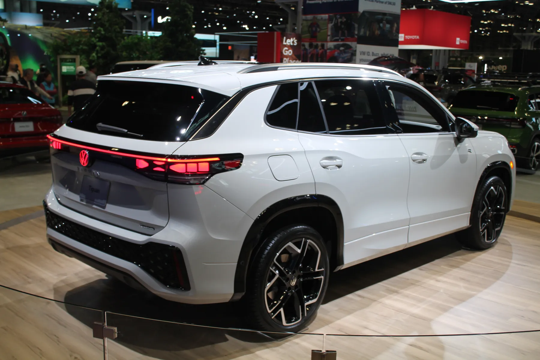 Rear three-quarter of a white Volkswagen Tiguan on a light wood auto-show floor, tail toward the left, flank running to the right, with a rope barrier in the foreground. A thin red lamp bar crosses the tail with a red illuminated VW roundel in the center and vertical red segments in the corner lamps. The lower bumper is black with a mesh section. The roof rails are silver. The wheel is black with bright machined edges on the spokes. A red car is at the left, a green SUV at the right, and a red Toyota sign hangs in the hall behind.