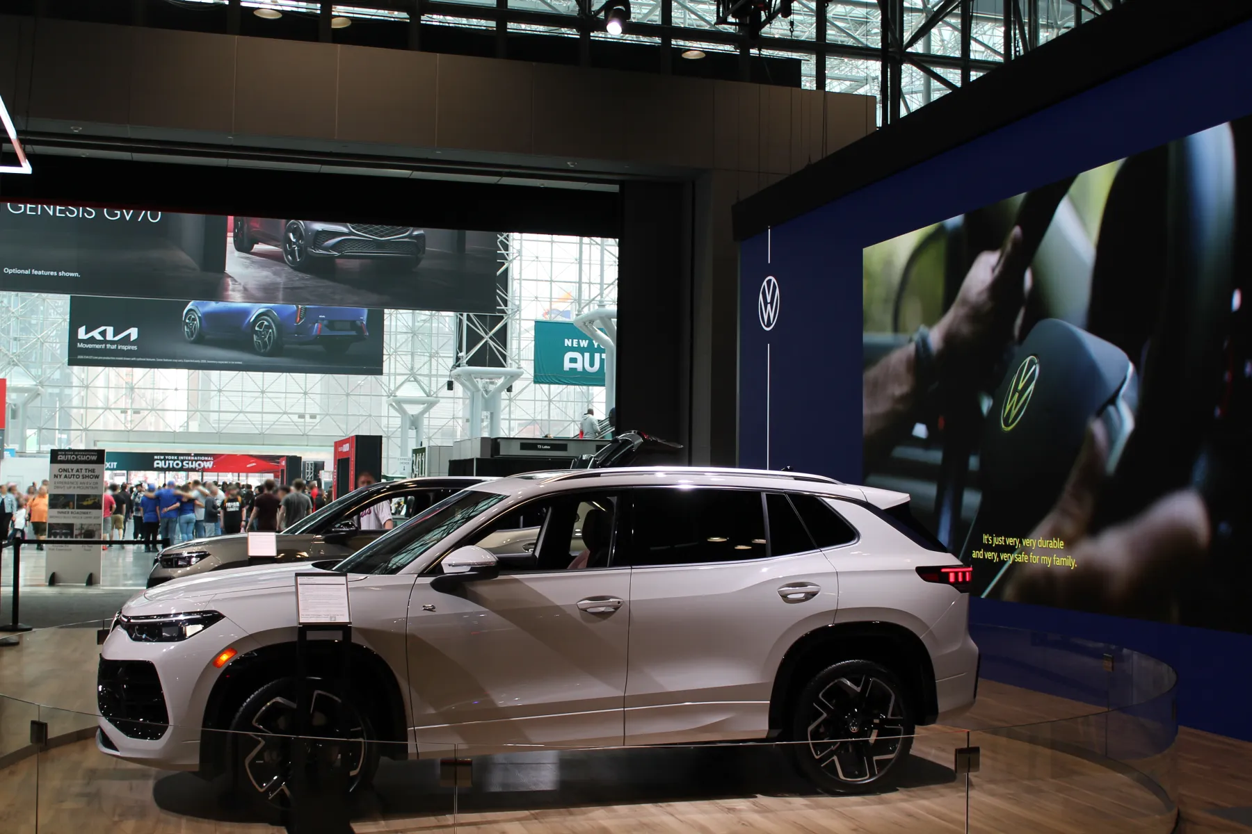 Side view of a white Volkswagen Tiguan on a round wood platform at an auto show, nose to the left, behind a glass barrier. Silver roof rails run above the windows. The mirror cap is white. The wheels are black with bright machined edges on the spokes. A white placard on a stand sits in front of the front wheel. Another light-colored SUV is parked behind it. A blue wall with a VW roundel stands to the right; its screen shows a hand on a VW steering wheel with yellow caption text. A crowd and other brand displays are farther back in the hall.