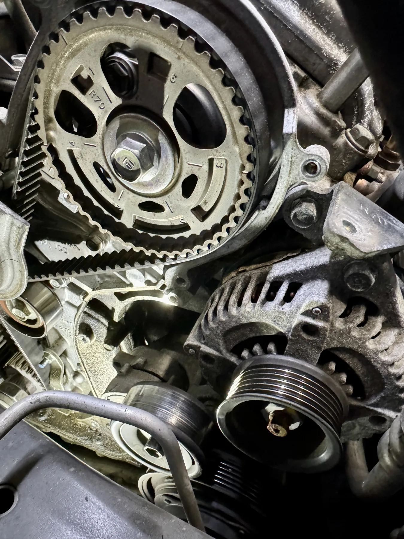 Close view in a dark engine bay, cover off the sprocket. A black toothed belt is meshed with a silver-gray metal sprocket. A hex bolt sits in the center. Stamped numerals on the sprocket, including 3, 4, and 6, and the characters R70 are stamped on the face. Another numeral is partly hidden. At the lower right a ribbed metal pulley sits in front of a finned housing.