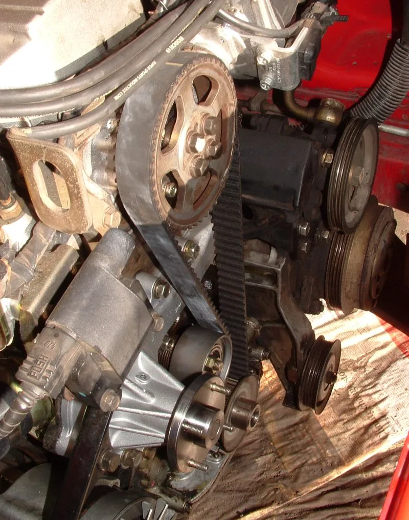Engine bay with the timing cover off. Red bodywork is at the right, and the ground is crumpled paper with print on it. A black toothed belt runs down the middle, meshed with a large rusty brown toothed metal sprocket at the top. Below the belt are a smooth silver idler pulley and a round hub with protruding studs. To the right are ribbed metal pulleys, one above another, and a smaller ribbed pulley near the bottom. Black ignition leads running across the top are printed BOSCH. At the lower left a gray housing has a connector marked PUSH.