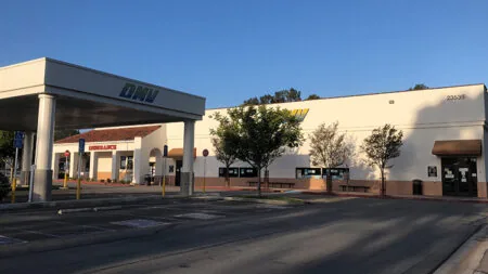 Low cream and white stucco building in late-afternoon light, a freestanding entrance canopy at the left with DMV in blue letters, a small red-brown tile roof over one storefront at the back left with a sign, the number 23535 high on the right wall, a few trees in front, an empty parking lot, and a clear blue sky