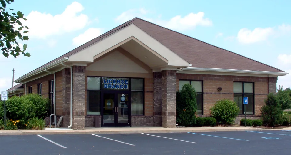 Single-story tan and brown brick building with a dark brown shingled hip roof and a cream gabled entry porch, blue letters reading LICENSE BRANCH on a white panel above the glass doors, 720 on the door glass, shrubs and a small evergreen in front, an empty parking lot with white and blue lines, blue sky with white clouds, and a tree branch at the upper left