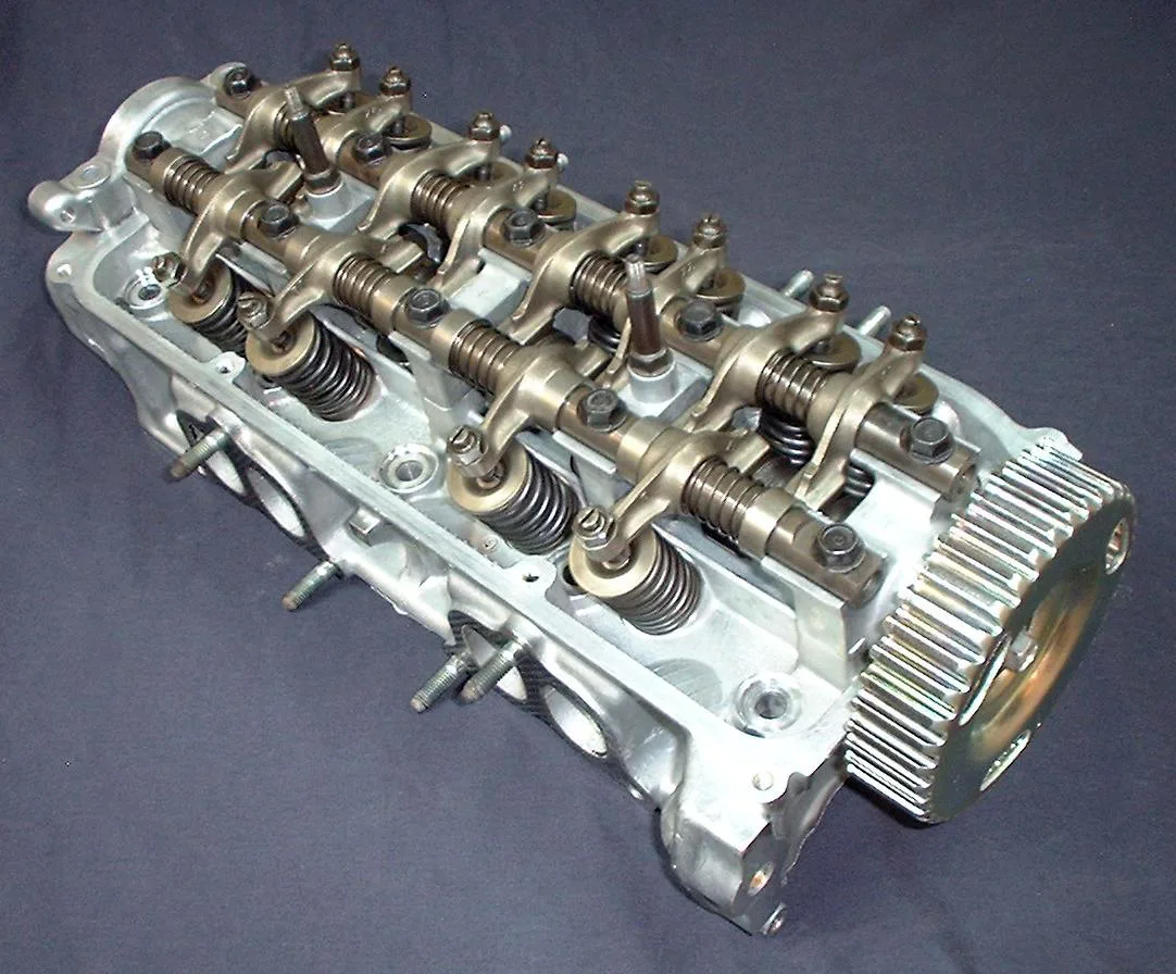 A silver cast-metal cylinder head on a dark blue cloth. Two shafts run the length of the head, carrying rocker arms with screws and nuts, and a large toothed pulley sits at the right end. Dark coil springs sit under the rocker arms. Threaded studs stick out of the near side, and round ports are open along the lower face. No printed text is visible.