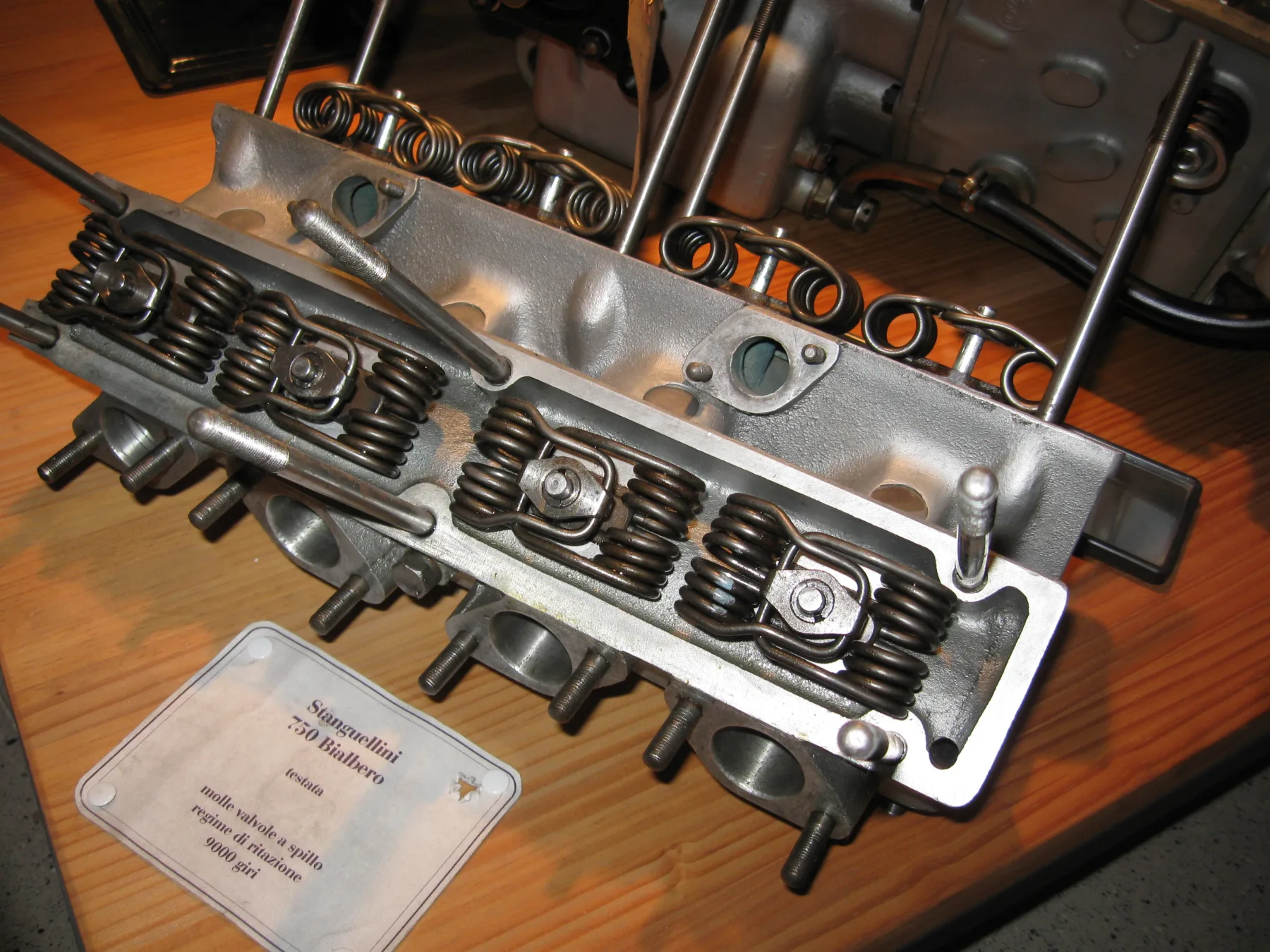 A cast-metal cylinder head on a light wooden table. Dark wire springs, each with several coils and long arms, are clamped under small plates and bolts. More of the same springs sit in a row along the far side. Long metal rods stand up from the head, and two lie across it. A card in front, held with white pins, includes the lines Stanguellini, 750 Bialbero, testata, molle valvole a spillo, and 9000 giri.