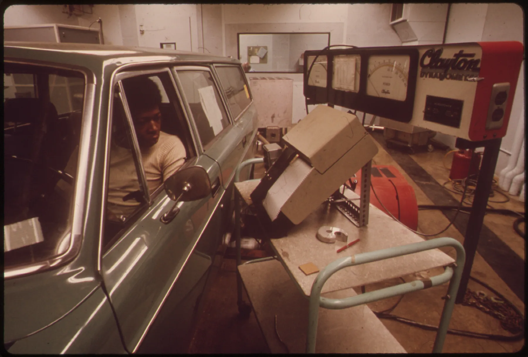 A 1970s color photograph. A pale green station wagon fills the left side, seen from the driver's side front quarter. A man in a cream sweater sits in the driver's seat, looking out the open window toward the side mirror. Beside the car, a beige boxy instrument housing sits tilted toward the driver on a metal cart with light-green tube railings. Behind it, a wall-mounted console has three large analog gauges and a red-trimmed panel lettered Clayton Dynamometer. Red equipment and cables lie on the floor at the right.