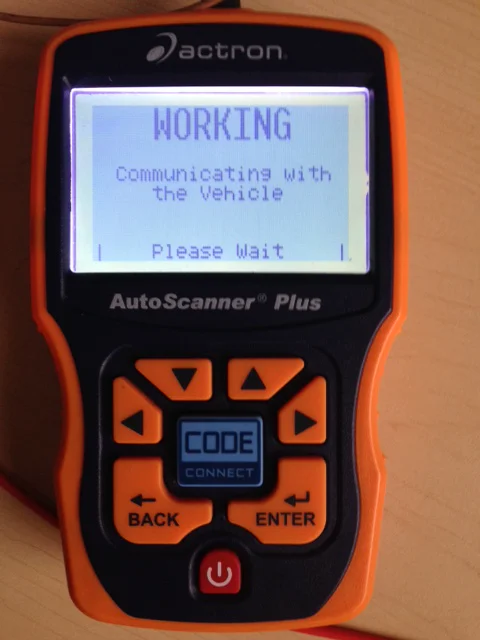 A close-up of an orange-and-black Actron AutoScanner Plus handheld code reader lying on a light wood surface. The screen reads WORKING, then Communicating with the Vehicle, then Please Wait. Orange arrow keys, a blue CODE CONNECT button, BACK and ENTER keys, and a red power button are on the face. A red cable enters at the bottom left edge.