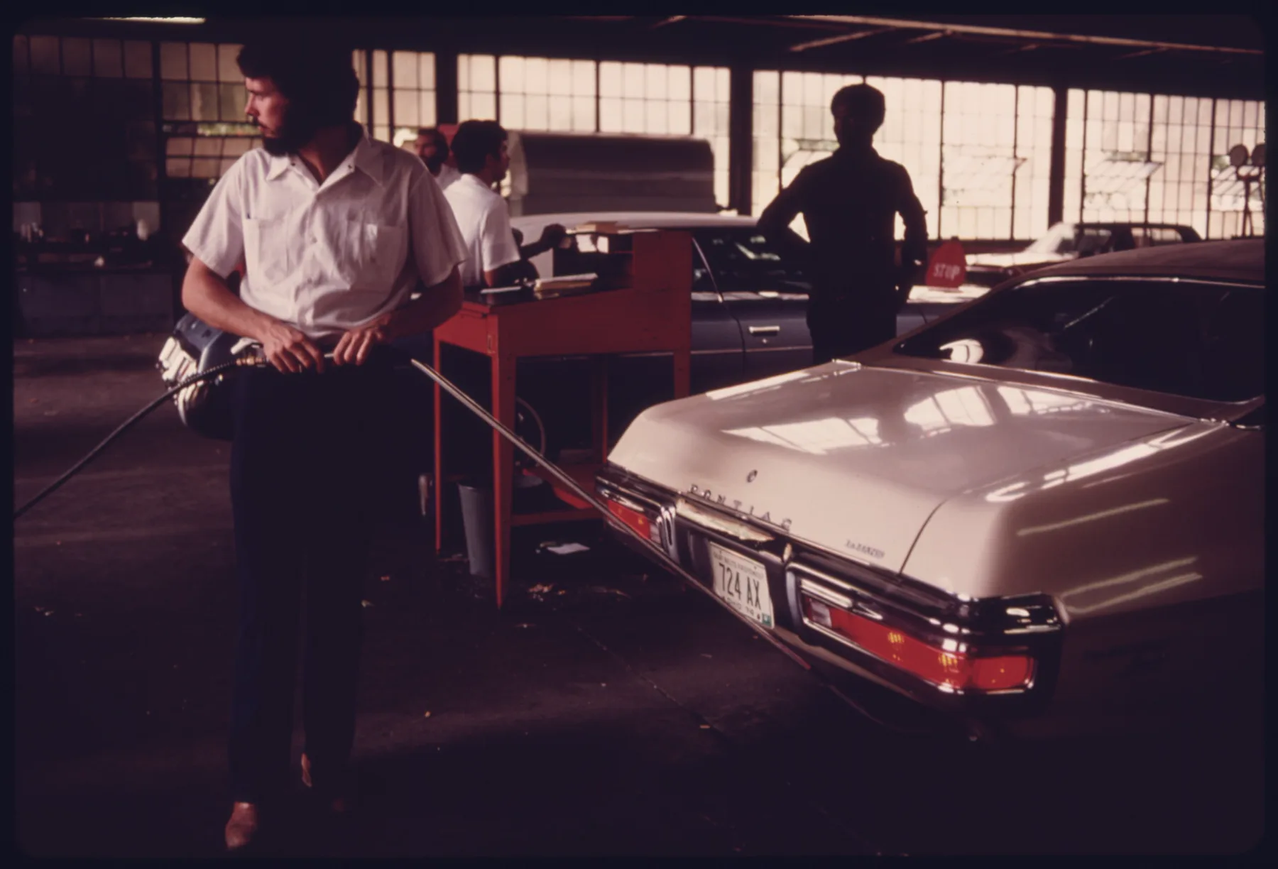 Inside a large inspection shed with tall windows, a bearded man in a white short-sleeved shirt and dark pants stands at the left holding a long metal probe on a hose that reaches toward the rear of a cream-white Pontiac sedan. The car is seen from the rear three-quarter, with a chrome bumper, a wide red horizontal taillamp, and a plate reading 724 AX. A red stand with papers is behind him. Other workers in white shirts and a dark silhouetted man stand farther back, with another car behind.