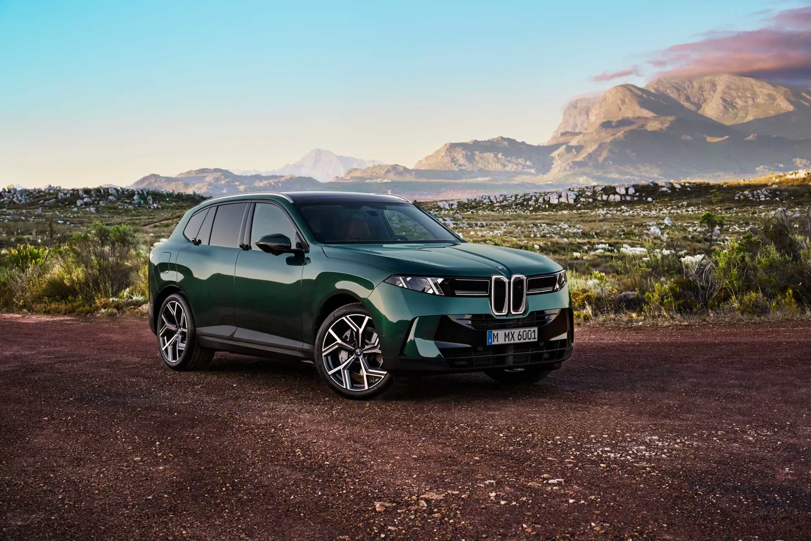 Green 2027 BMW X5, front three-quarter on a mountain path with double-X lamps and vertical kidney Iconic Glow