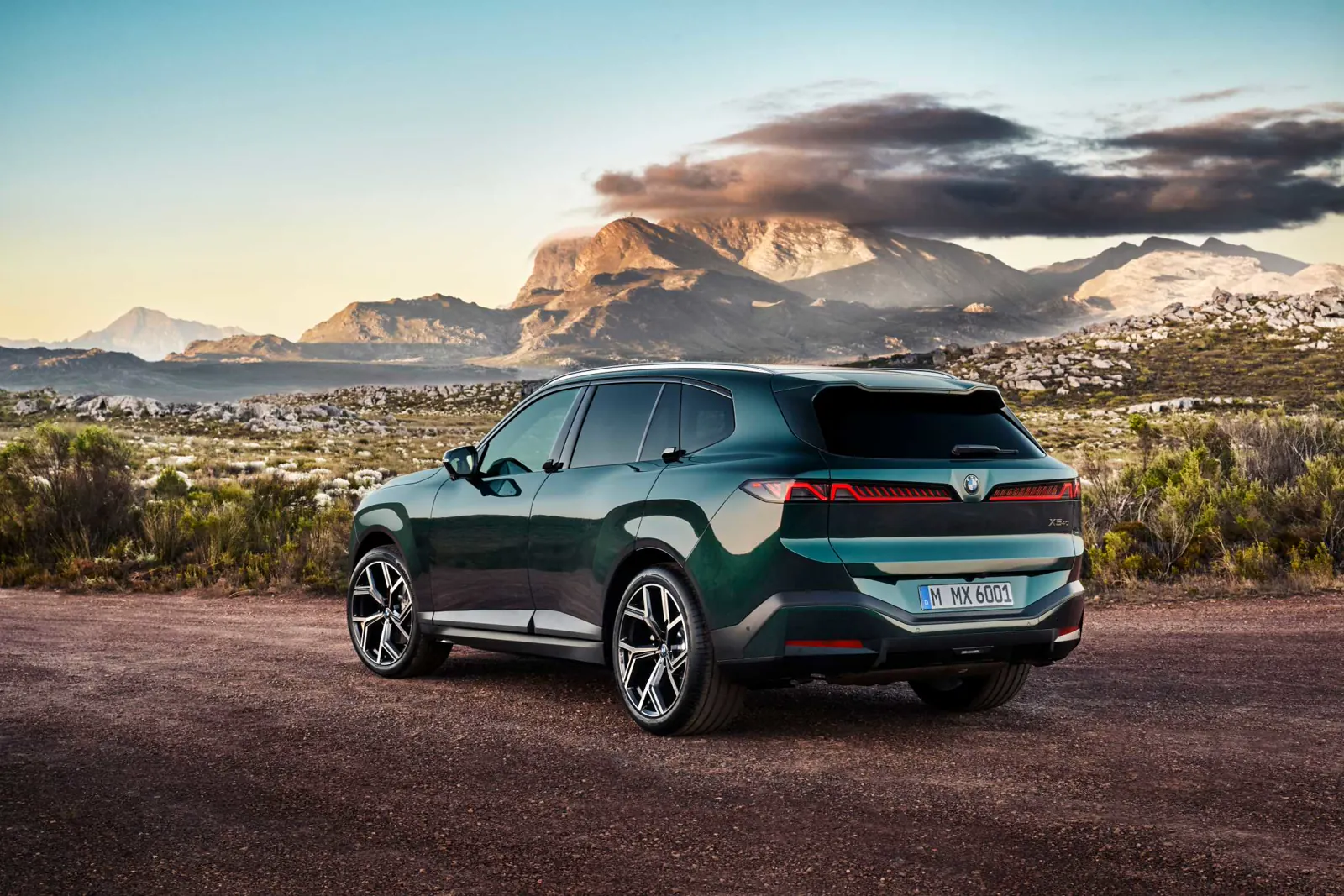Green 2027 BMW X5, rear three-quarter on a mountain path with slim lamps echoing the double-X